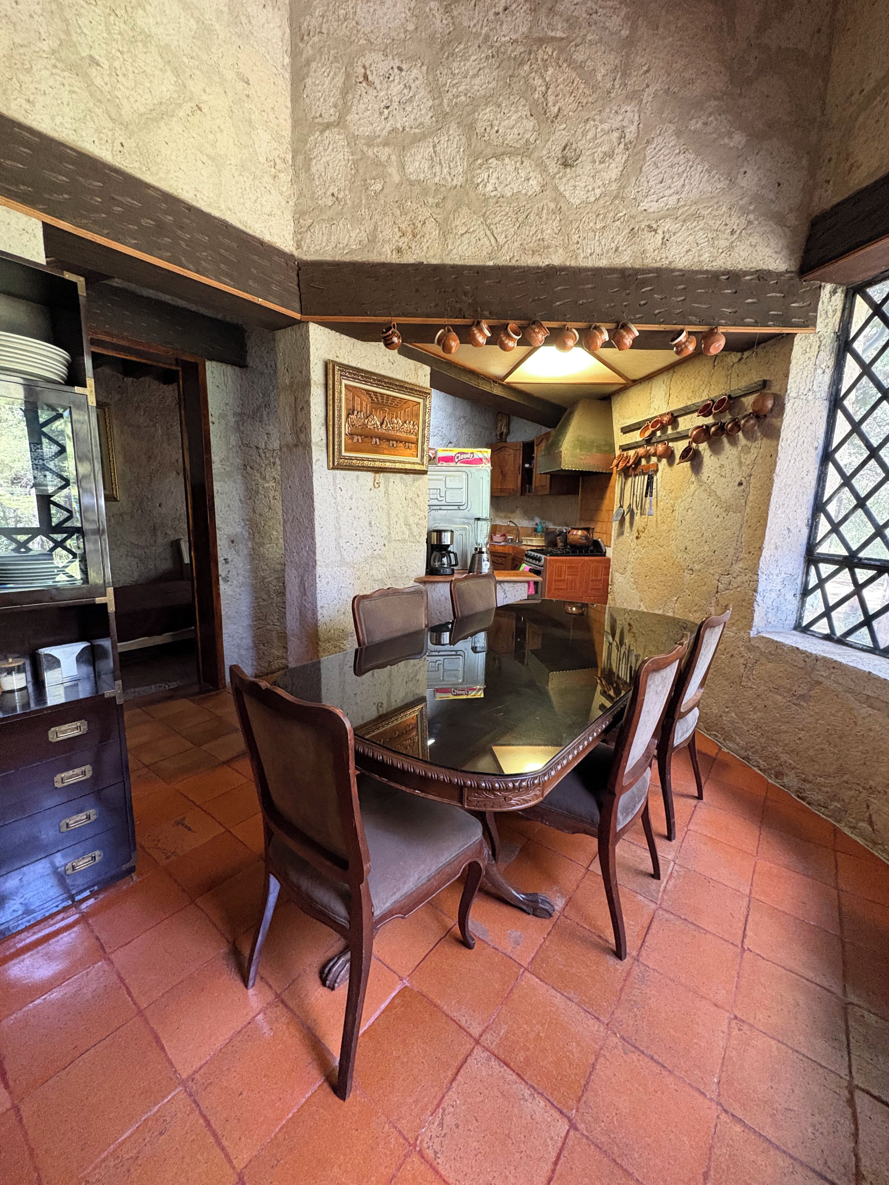A dining area with a glass table surrounded by chairs, a kitchen in the background, and a window with a lattice design.