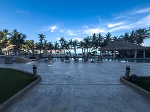 Ein Poolbereich eines Resorts mit Liegestühlen, einer Struktur mit Reetdach und Palmen.