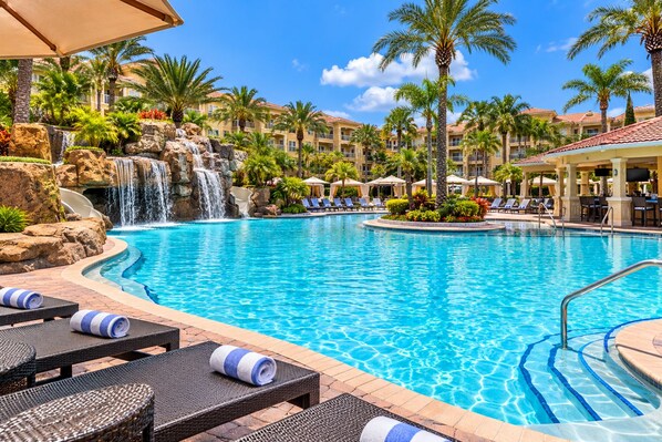 Piscina de resort con cascada, sillas de descanso y palmeras.