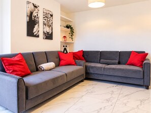Ein modernes Wohnzimmer mit einem grauen Ecksofa, roten Kissen, einem weißen Boden und Bildern an der Wand.