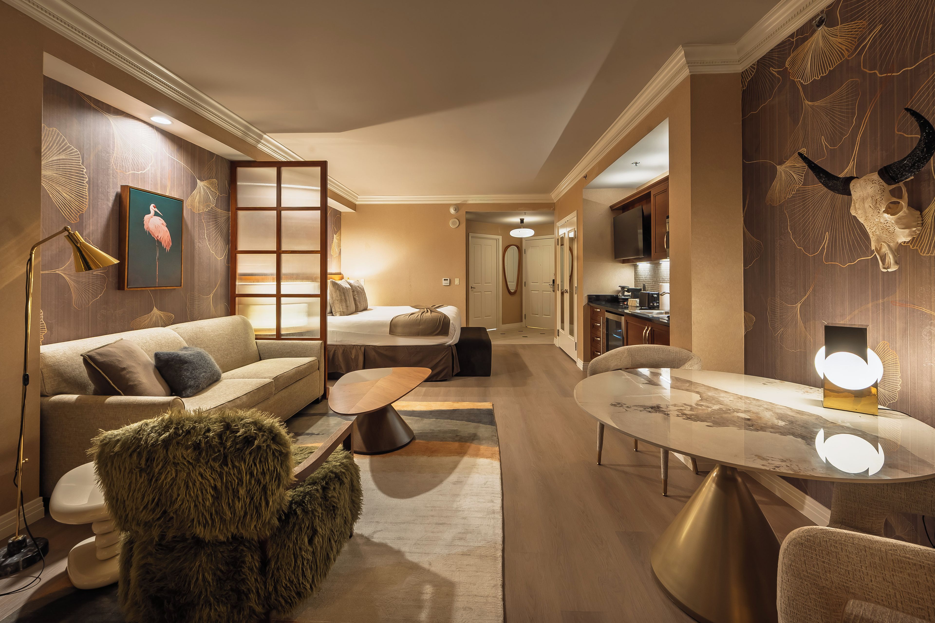 A hotel room with a sofa, a coffee table, a dining table, a lamp, a painting, and a decorative wall.