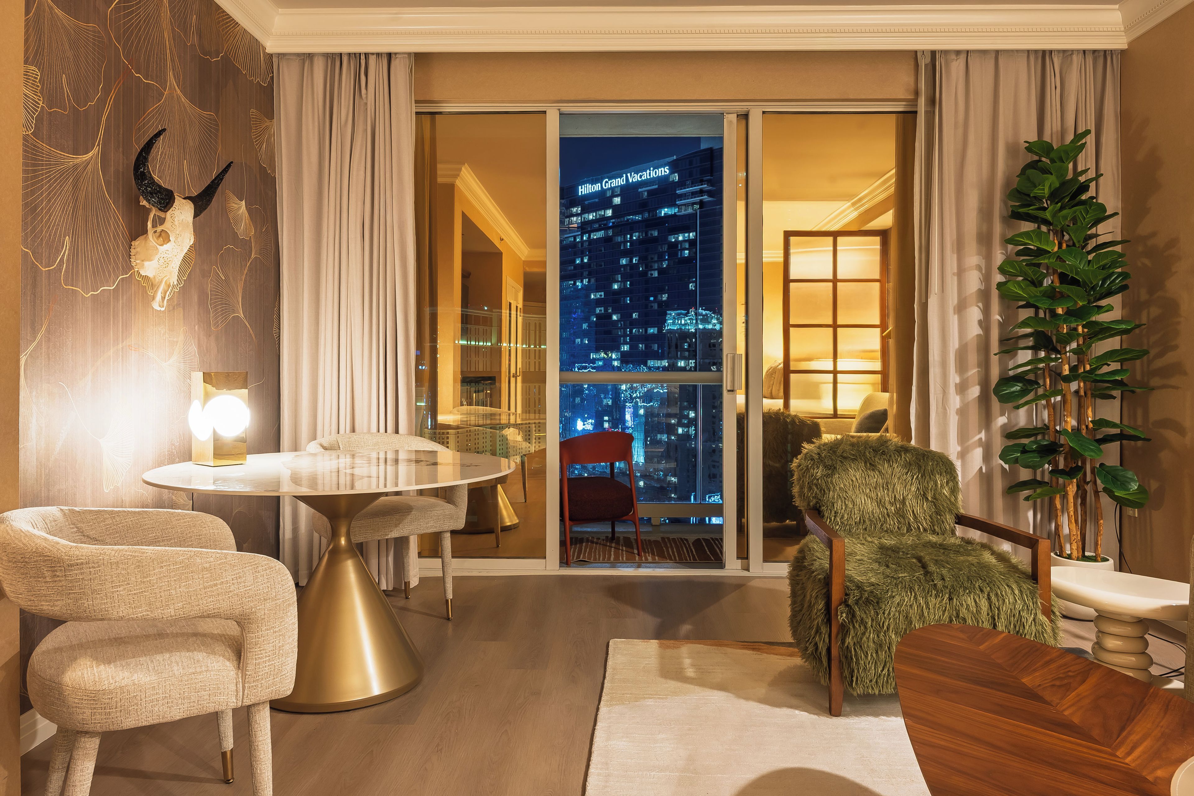 A modern hotel room with a city view, a round table, two chairs, and a large potted plant.