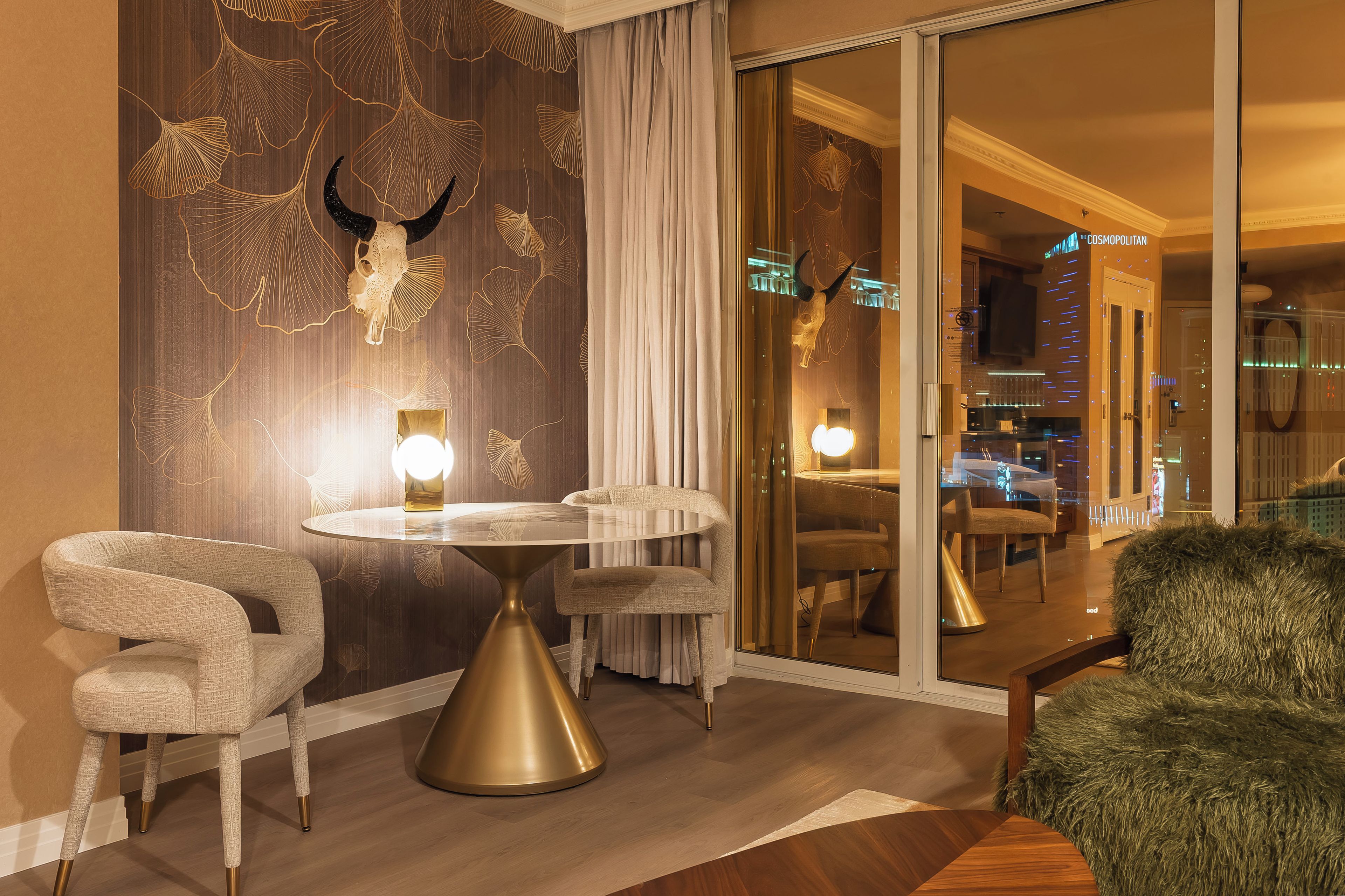 Une chambre d’hôtel moderne avec une table ronde, deux chaises, un plaid en fausse fourrure et un mur décoratif orné d’un motif représentant un crâne et des feuilles de ginkgo.