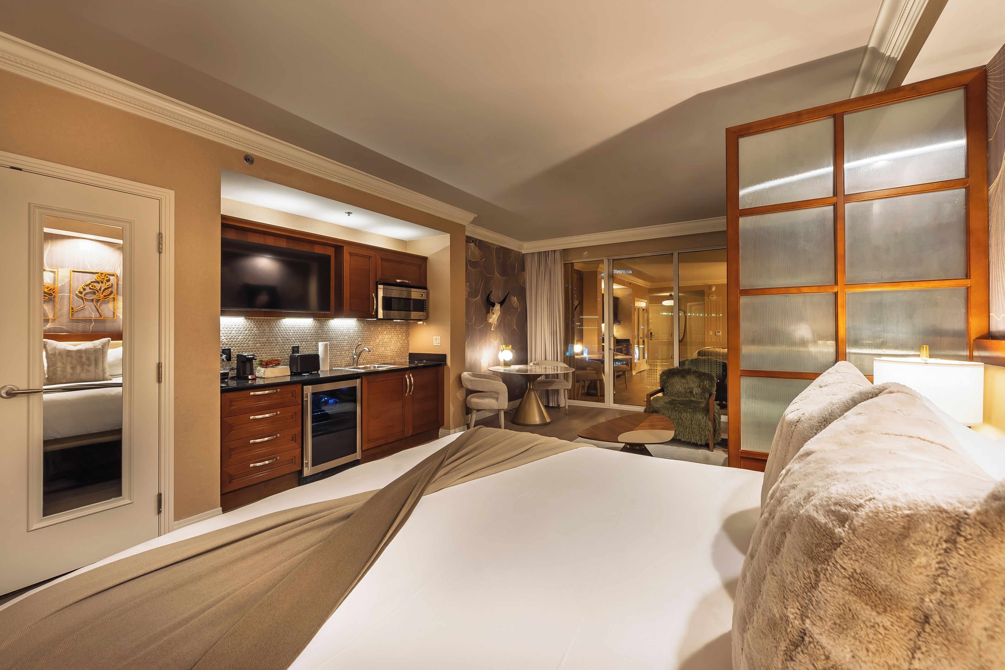 A hotel room with a kitchenette, a bed, a sitting area, and a view of the hotel lobby.