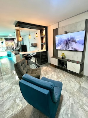Uma sala de estar moderna com uma TV de tela plana, um sofá e um espaço de jantar com cozinha ao fundo.