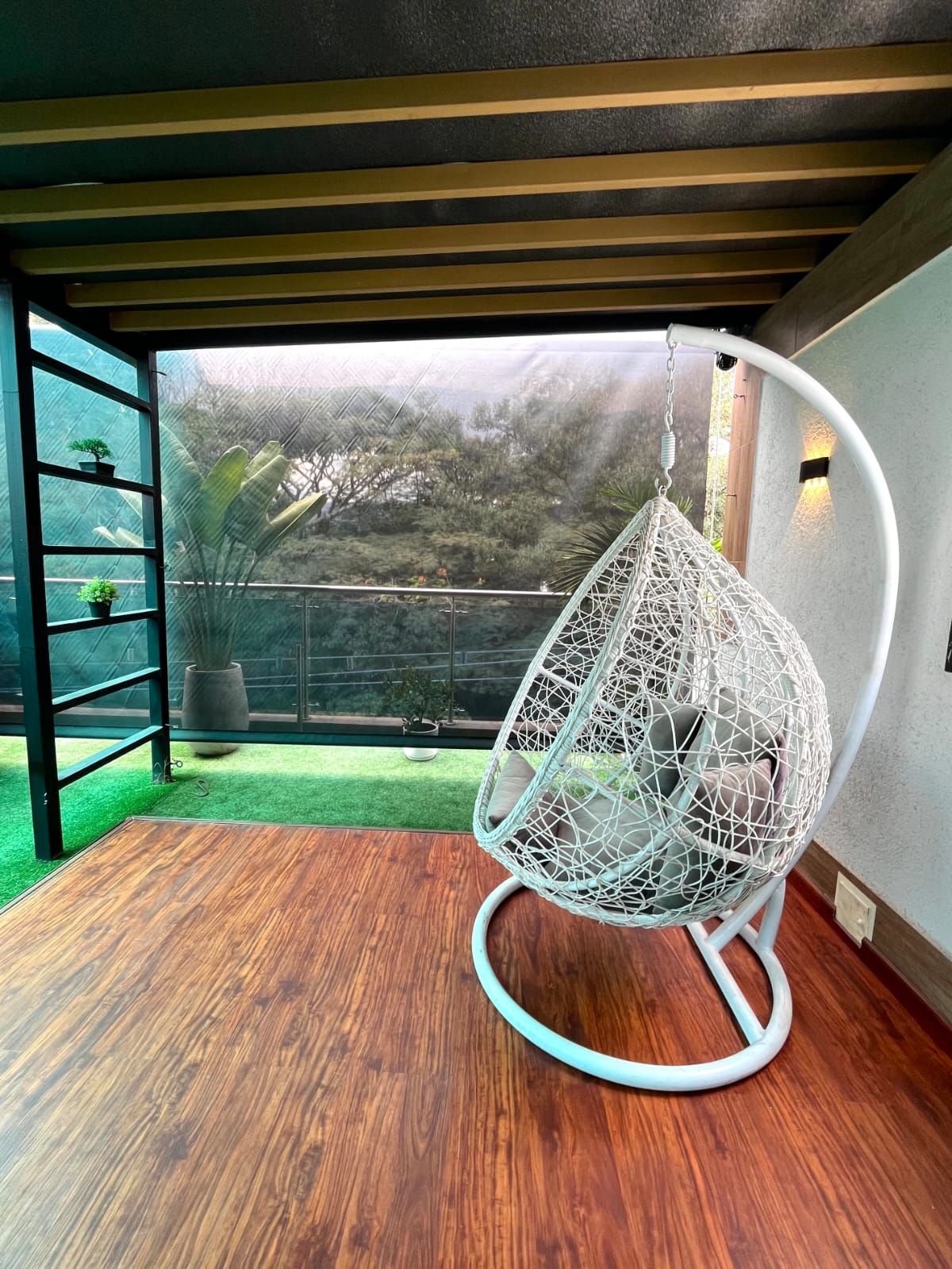 Quarto com cadeira suspensa, piso de madeira, porta de vidro que dá para um jardim e guarda-corrimão de metal.