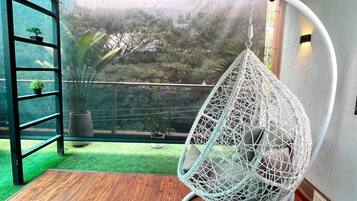 A room with a hanging chair, a wooden floor, a glass door leading to a garden, and a metal railing.