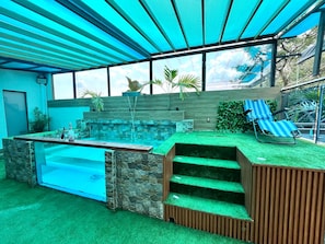 Uma área de piscina moderna ao ar livre, com parede de pedra, cascata pequena e área de estar com espreguiçadeira listrada em azul.