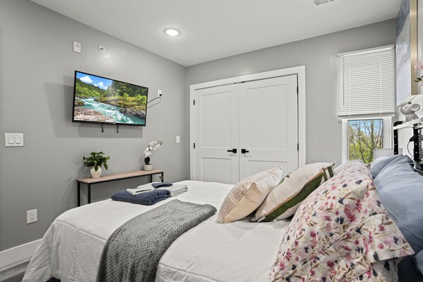 A bedroom with a bed, a TV mounted on the wall, a bedside table with a plant, and a window with blinds.