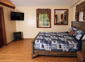Une chambre avec un plancher en bois, un lit recouvert d’une courtepointe à motifs, un téléviseur fixé au mur et une fenêtre avec des rideaux.