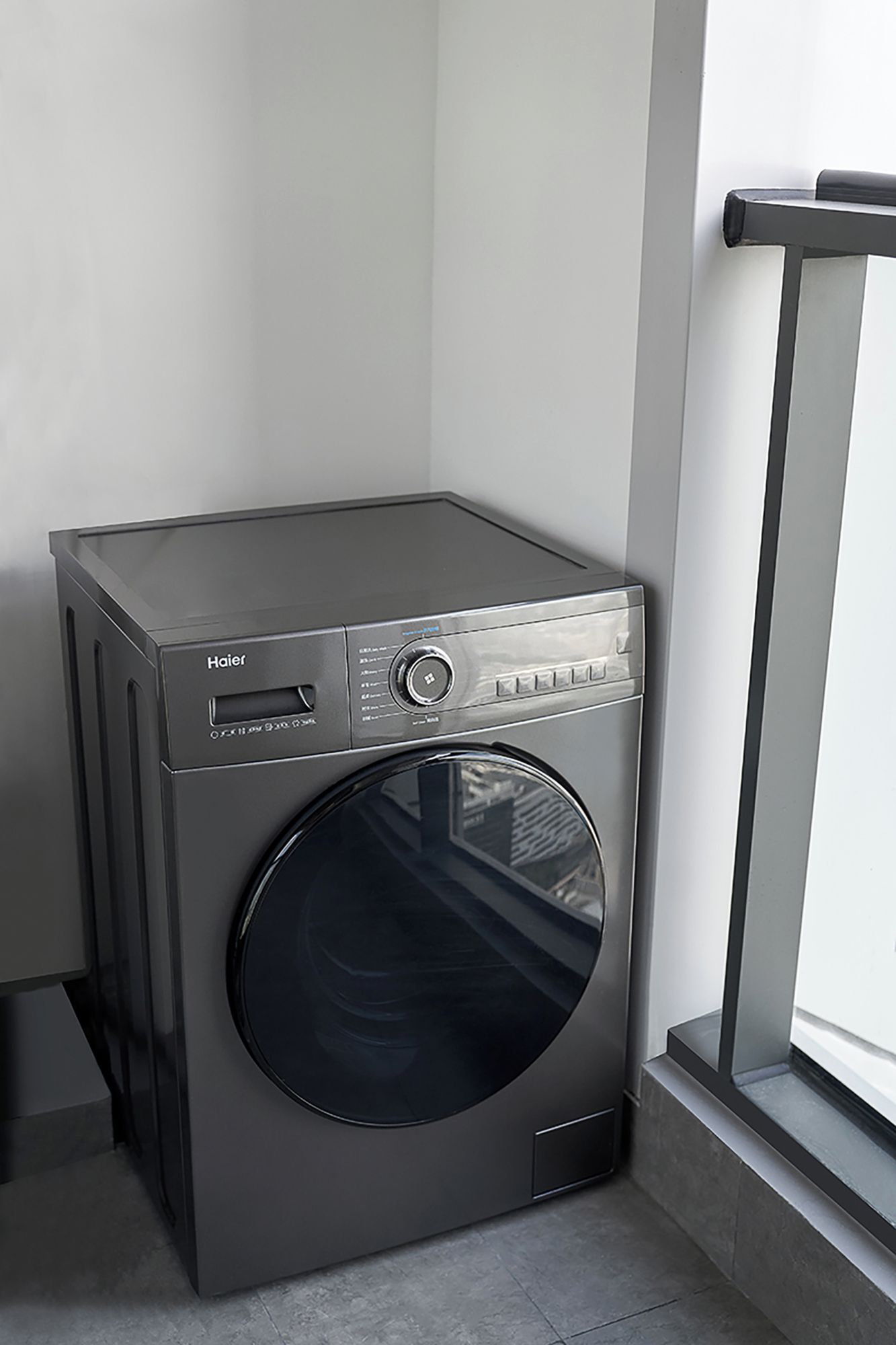 A compact, modern front-loading Haier washing machine in a corner of a room with a window.