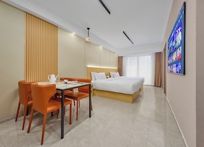 A hotel room with a bed, a dining table with chairs, a coffee table, a TV on the wall, and a large window.
