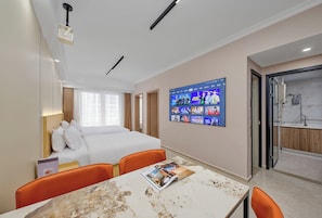 Habitació d'hotel amb llit, televisió penjada a la paret, taula amb una revista i un bany visible a través d'una porta oberta.