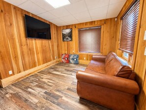 Ein Zimmer mit Holzwänden, einem Ledersofa, einem Flachbildfernseher und einem Fenster mit Jalousien.