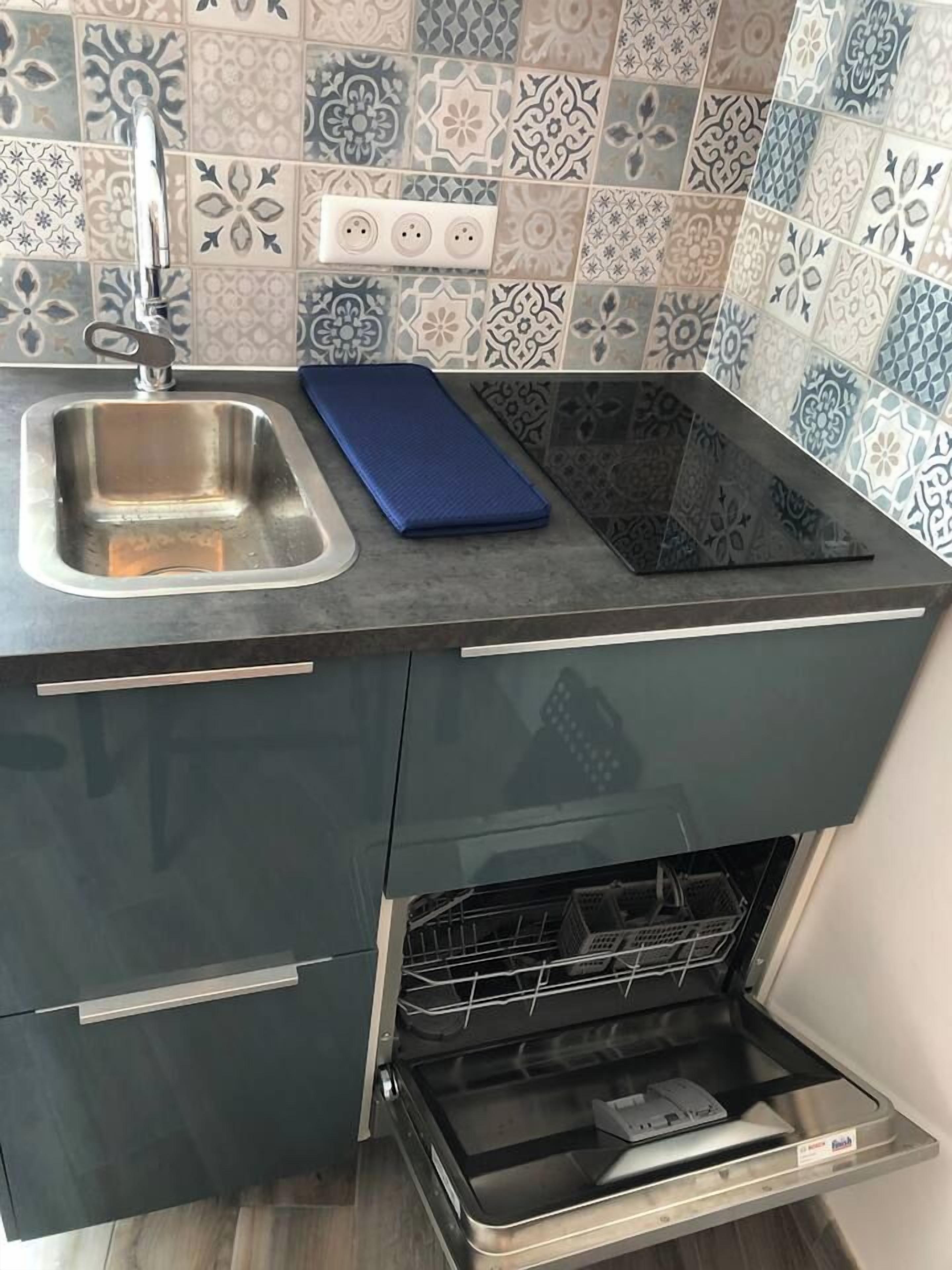 Una cocina con lavavajillas empotrado, un lavamanos de acero inoxidable y una tabla de cortar azul sobre la encimera.
