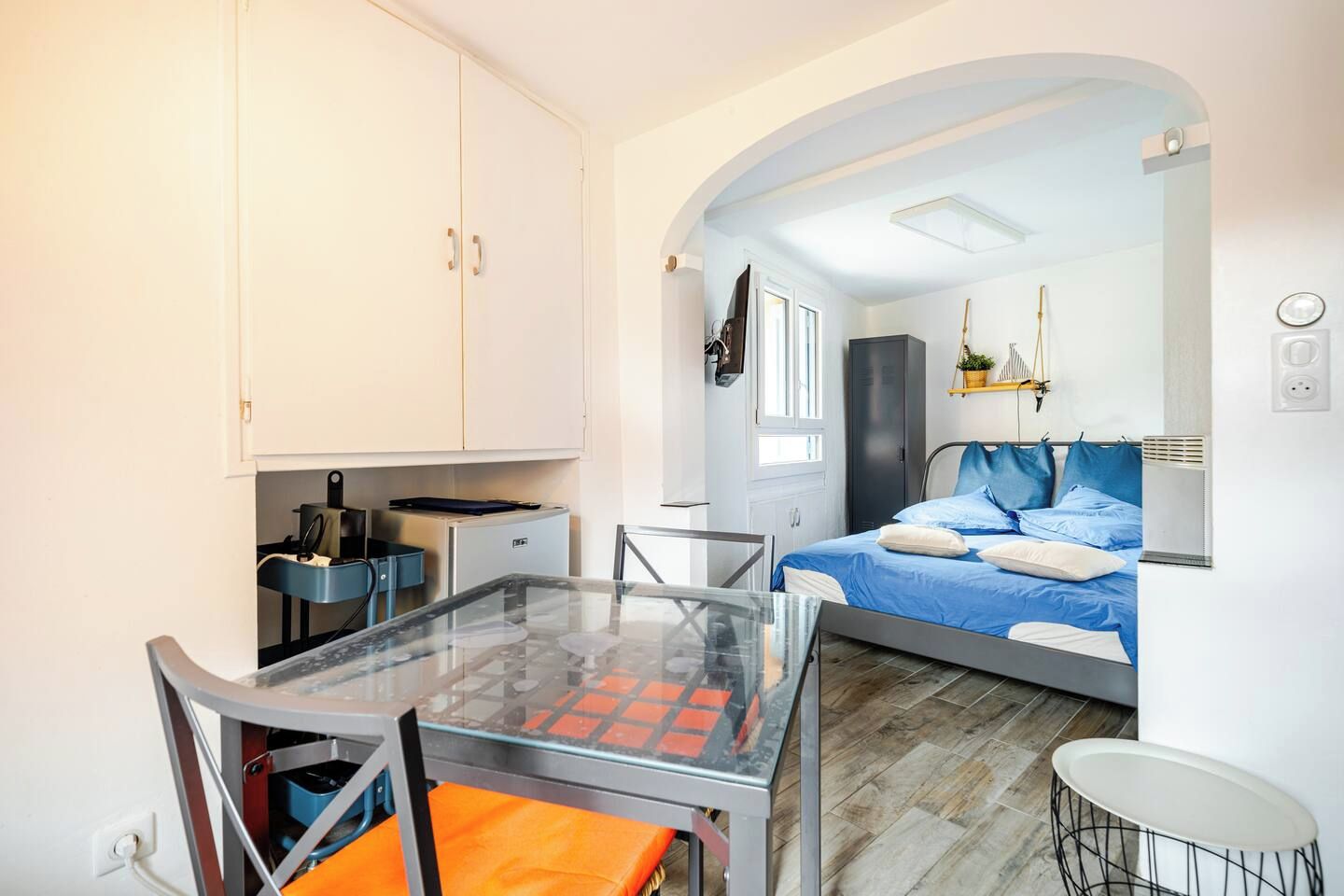 Une pièce compacte comprenant une table en verre, un lit avec une literie bleue, une kitchenette et une petite salle de bain.