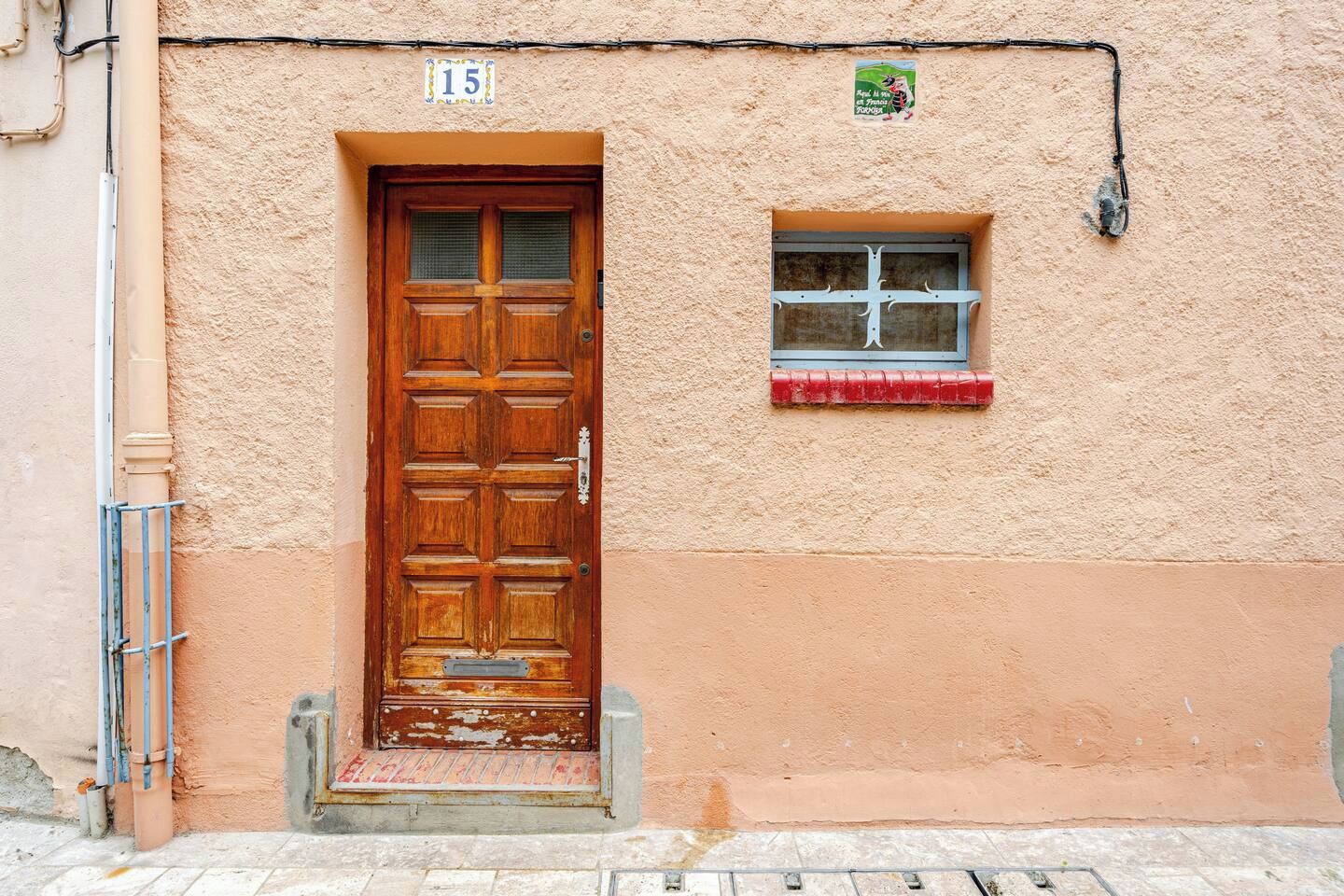 Una puerta de madera con una placa de número, un pequeño ventanal con marco metálico y un felpudo rojo en la entrada.