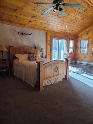 A bedroom with a wooden ceiling, a bed with a headboard, a nightstand, a lamp, and a window. - Larch Ridge Ranch Upper Unit (Arkport)
