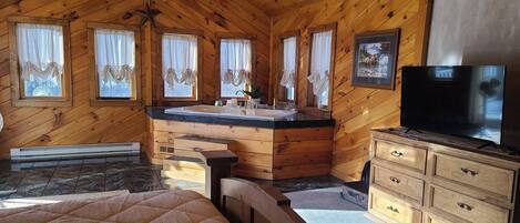A cabin interior with a wooden ceiling, a large bed, a bathroom with a sink and vanity, and a television.