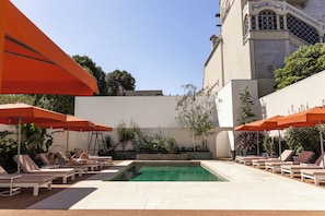 Área de piscina com espreguiçadeiras e guarda-sóis, cercada por um muro baixo e um edifício com detalhes decorativos.