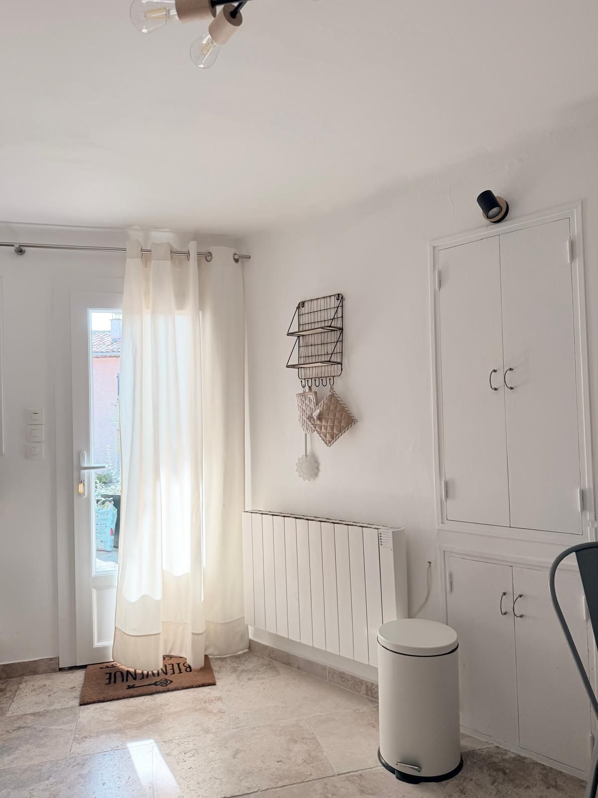 A room with a radiator, a white door, a radiator cover, a white trash bin, and a chair.