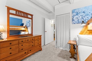 Ein Schlafzimmer mit einem Holzkasten, einem Bett mit weißer Bettwäsche, einem Blumenbild, einer Lampe und einer Tür zu einem anderen Raum.