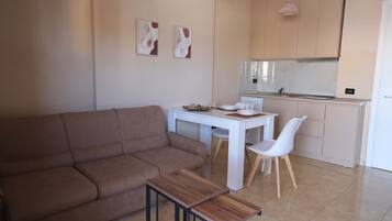Un salon moderne avec un canapé marron, une table à manger blanche et un coin cuisine doté d’un réfrigérateur blanc et de placards.