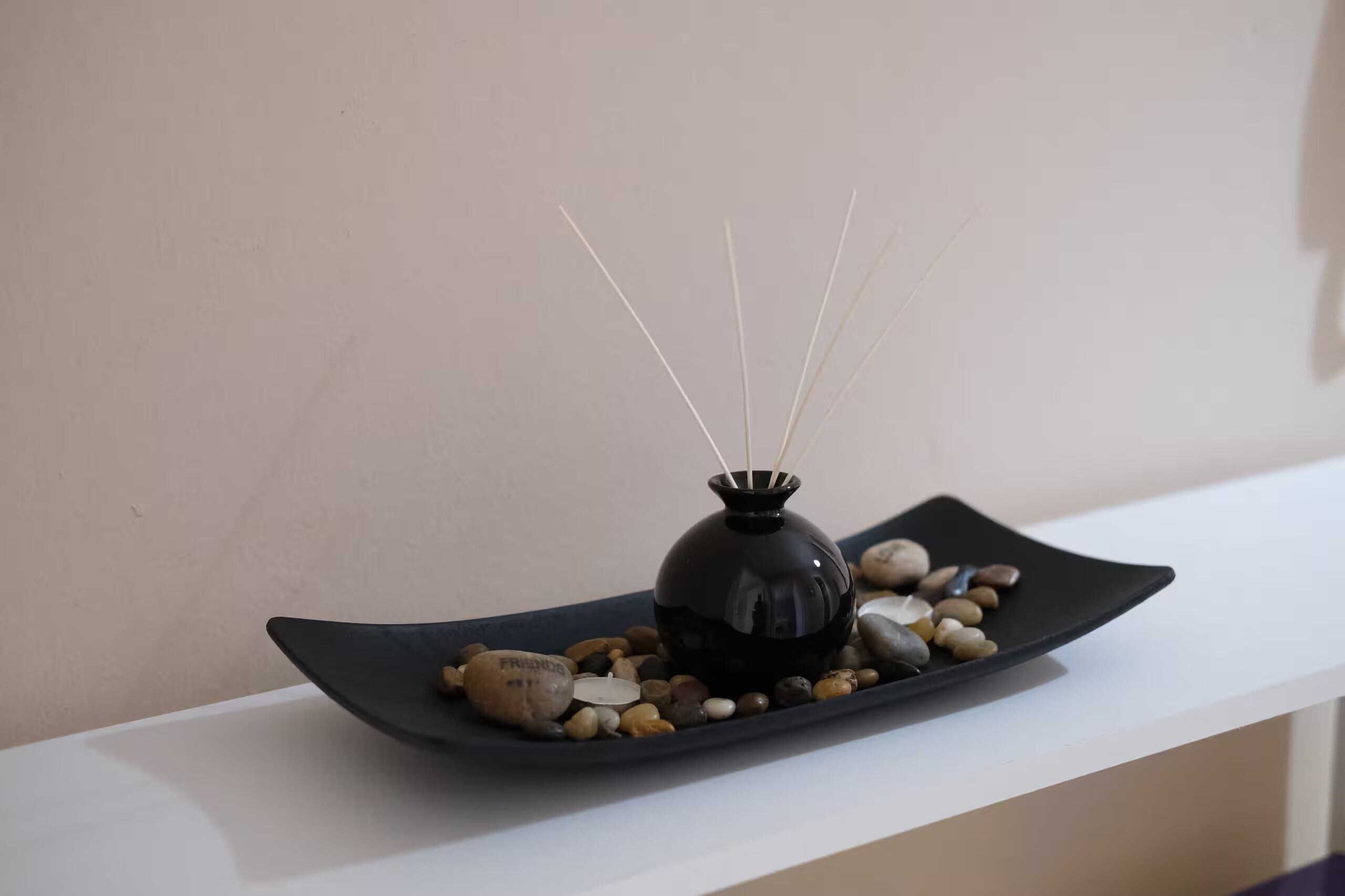 A black vase with reed sticks on a black plate with pebbles, placed on a white shelf.