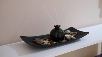 A black vase with reed sticks on a black plate with pebbles, placed on a white shelf.