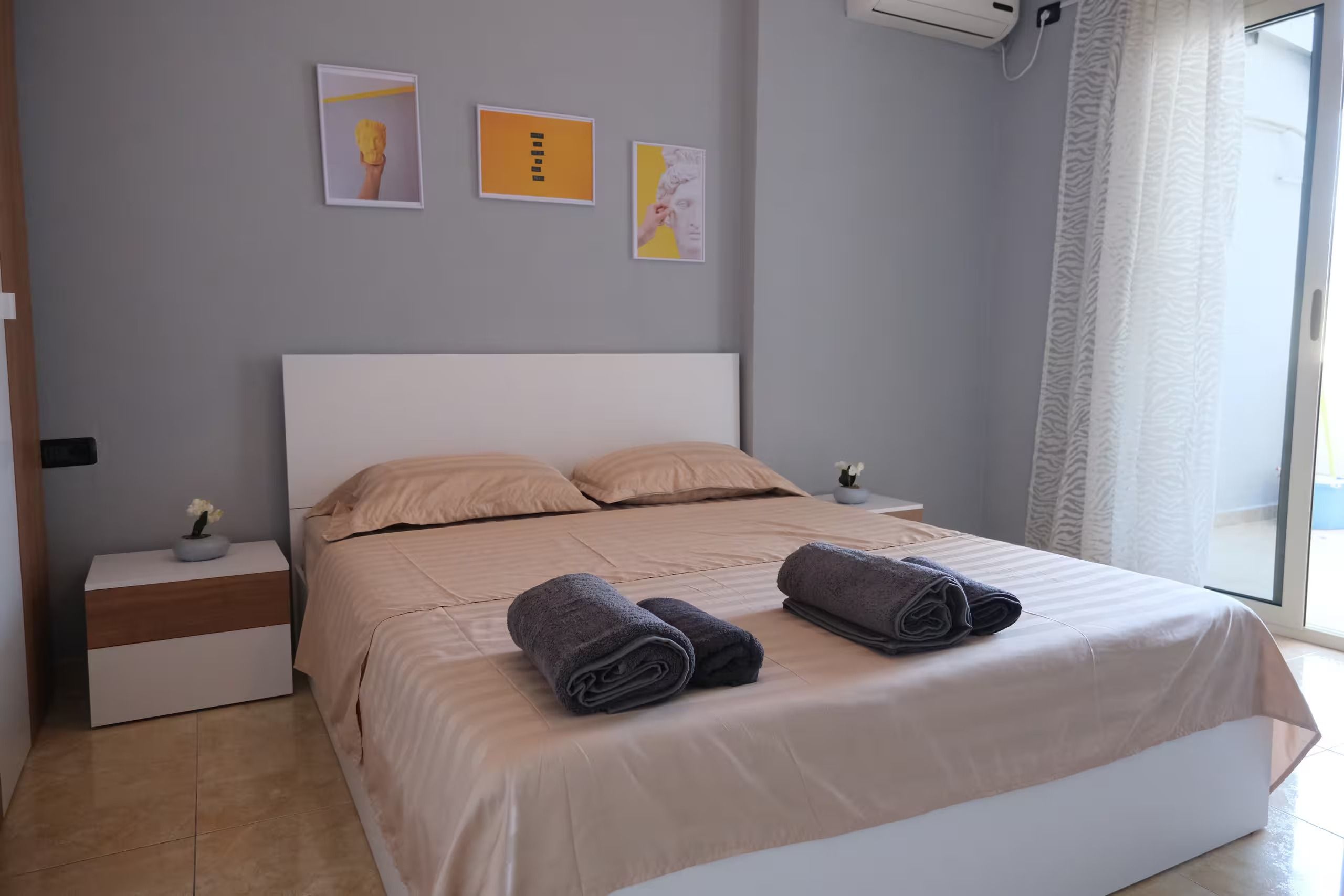 Une chambre à coucher avec un lit, deux tables de chevet, une fenêtre avec des rideaux et des œuvres d’art murales.