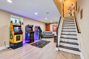A living room with arcade games, a grey sofa, a patterned rug, and a staircase. - Cape Cod Style 5-Bedroom Retreat with a wrap-around Porch in Vibrant Baltimore (Baltimore)