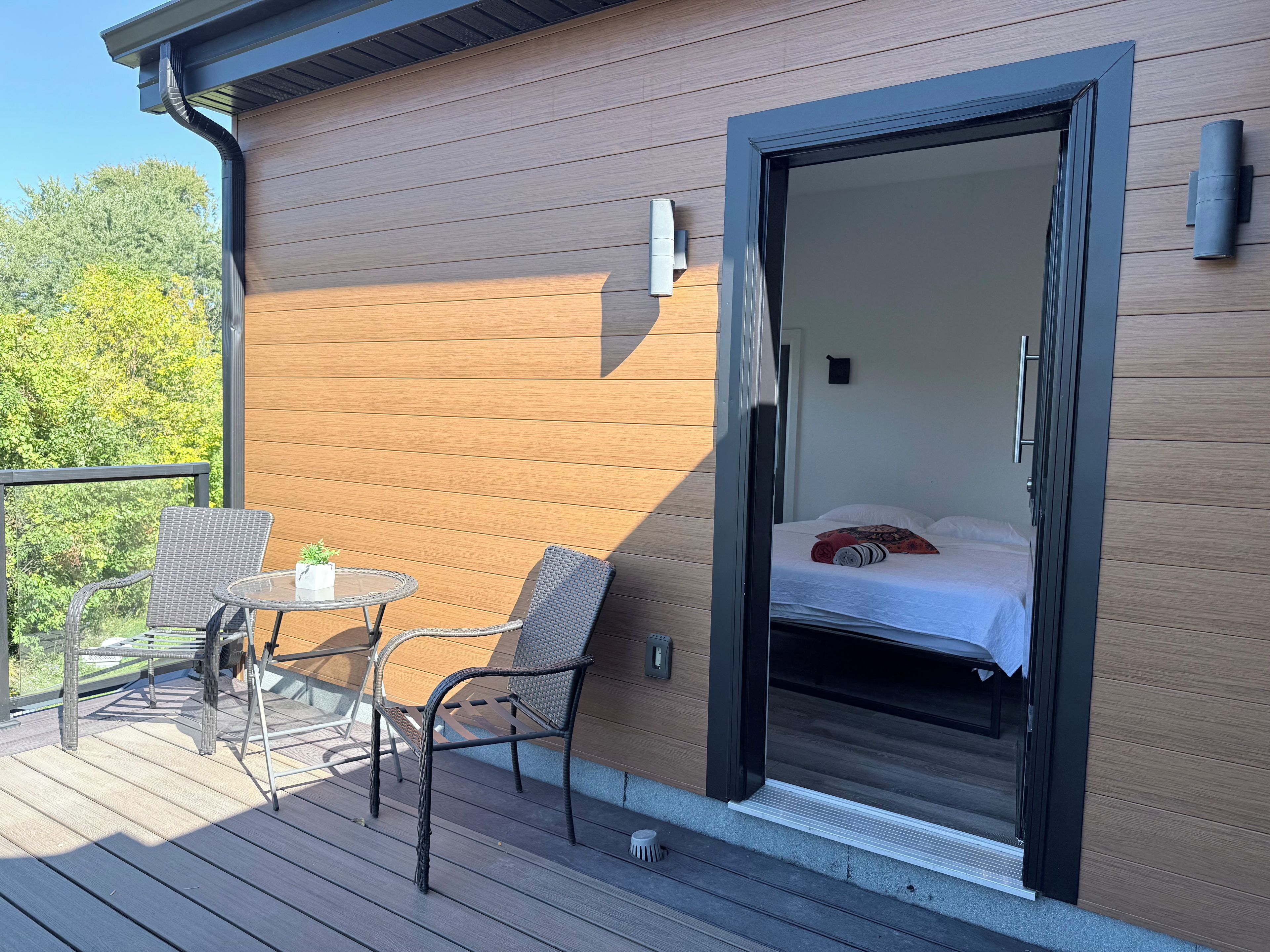 En balkon med fletstole og et lille bord, en skydedør til et soveværelse med seng og puder, samt en ydre væg i træ.