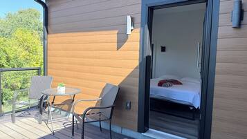 Een balkon met rieten stoelen en een tafeltje, een schuifdeur naar een slaapkamer met een bed en kussens, en een houten buitenmuur.