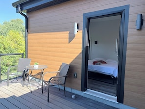 Ein Balkon mit Korbstühlen und einem kleinen Tisch, eine Schiebetür zu einem Schlafzimmer mit Bett und Kissen, sowie eine Außenwand aus Holz.