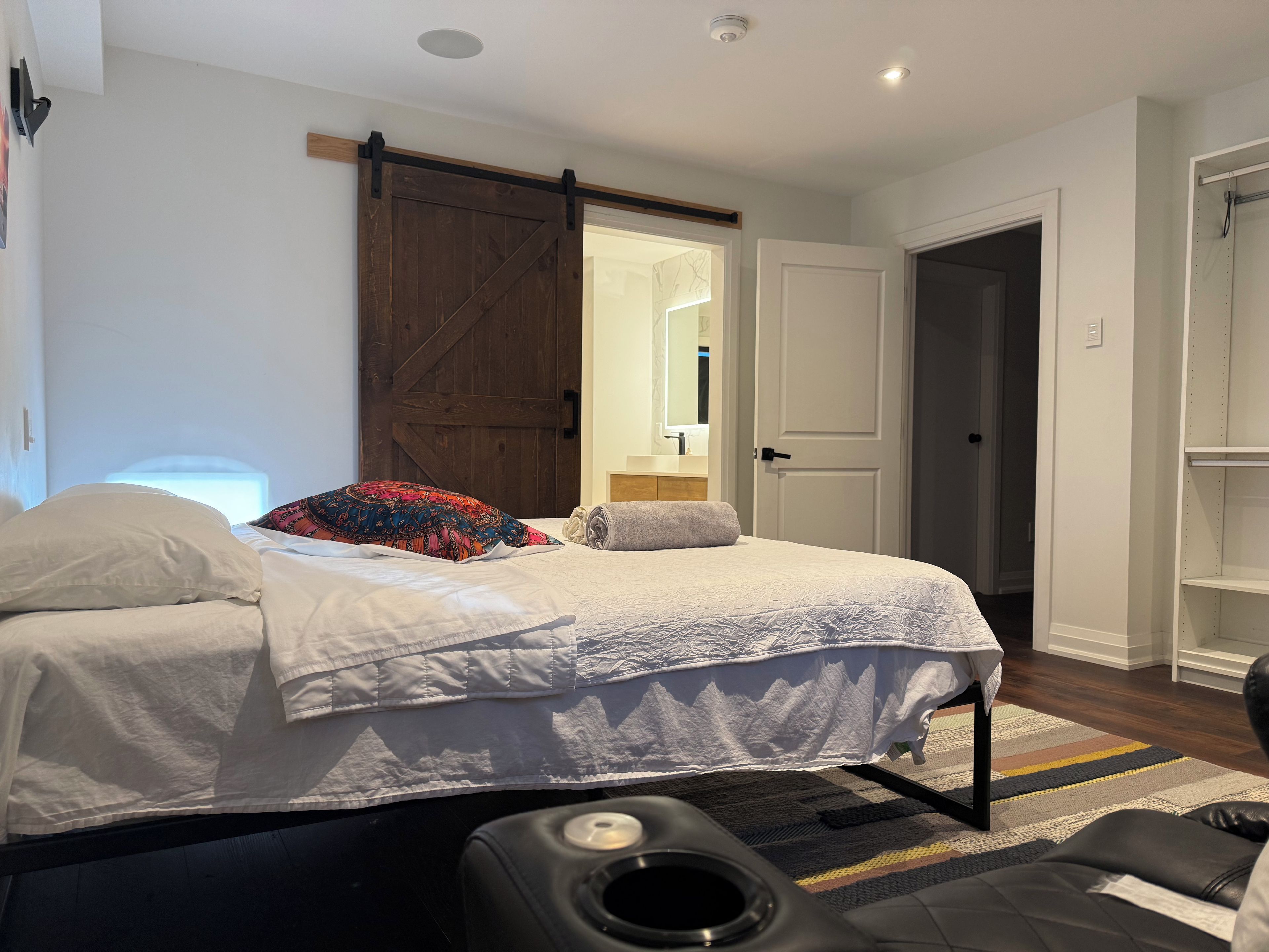 Un dormitorio con cama, un espejo grande, una puerta corrediza tipo granero y un reposapiés negro.
