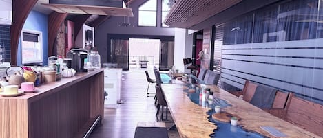 Una cocina moderna con una barra de madera alargada, un comedor con mesa de vidrio y vistas al exterior.