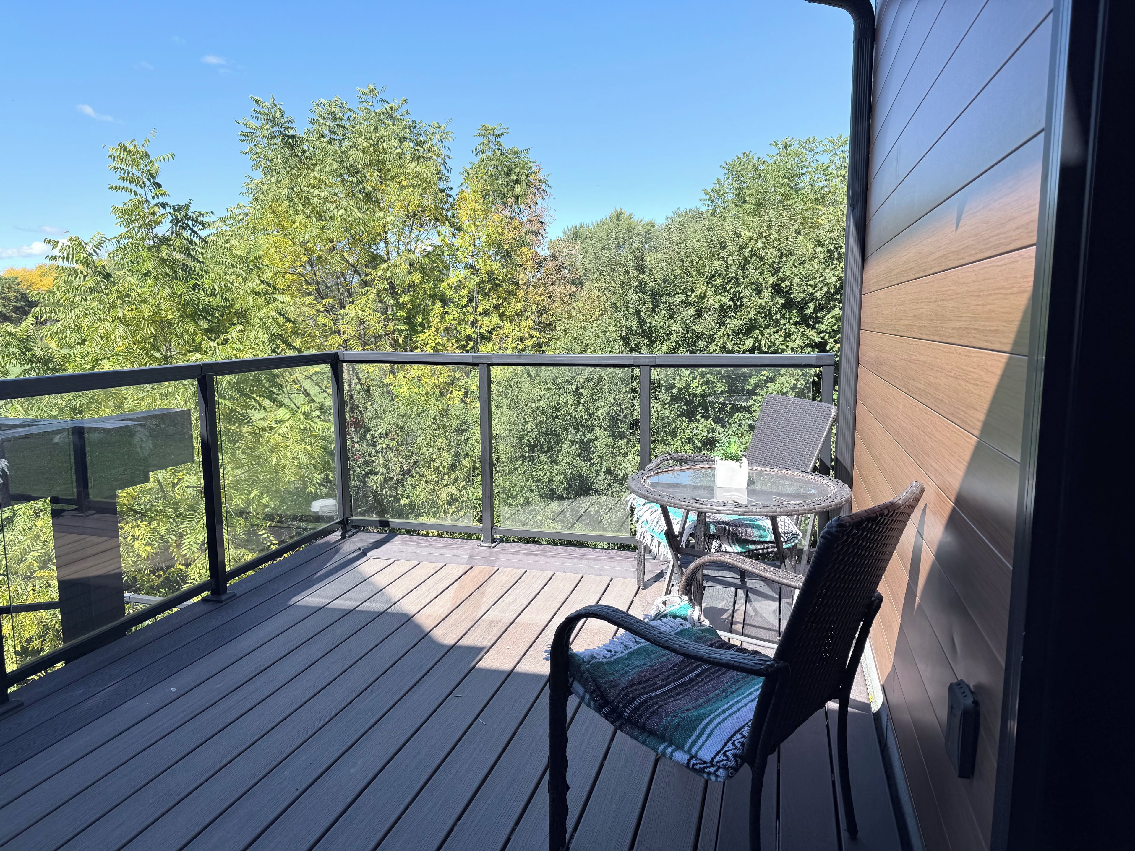 A wooden deck with a glass balustrade, wicker furniture, and a view of trees.