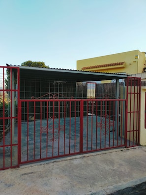A gated area with a metal fence, a covered section, and a building with a yellow facade in the background. - A bungalow perfect for families. Air conditioning in every room. Access to the pool. (L'Alfàs del Pi)