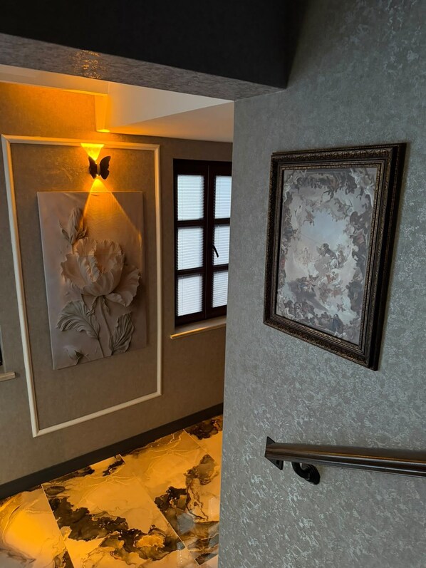 A hallway with a wall-mounted light fixture, a framed artwork, and a window with blinds. - SUR TERAS OTEL (Diyarbakir)