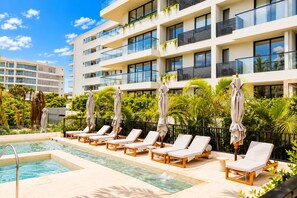 Un espace piscine d’hôtel avec des chaises longues et des parasols, entouré d’une végétation luxuriante et agrémenté d’une façade de bâtiment moderne.