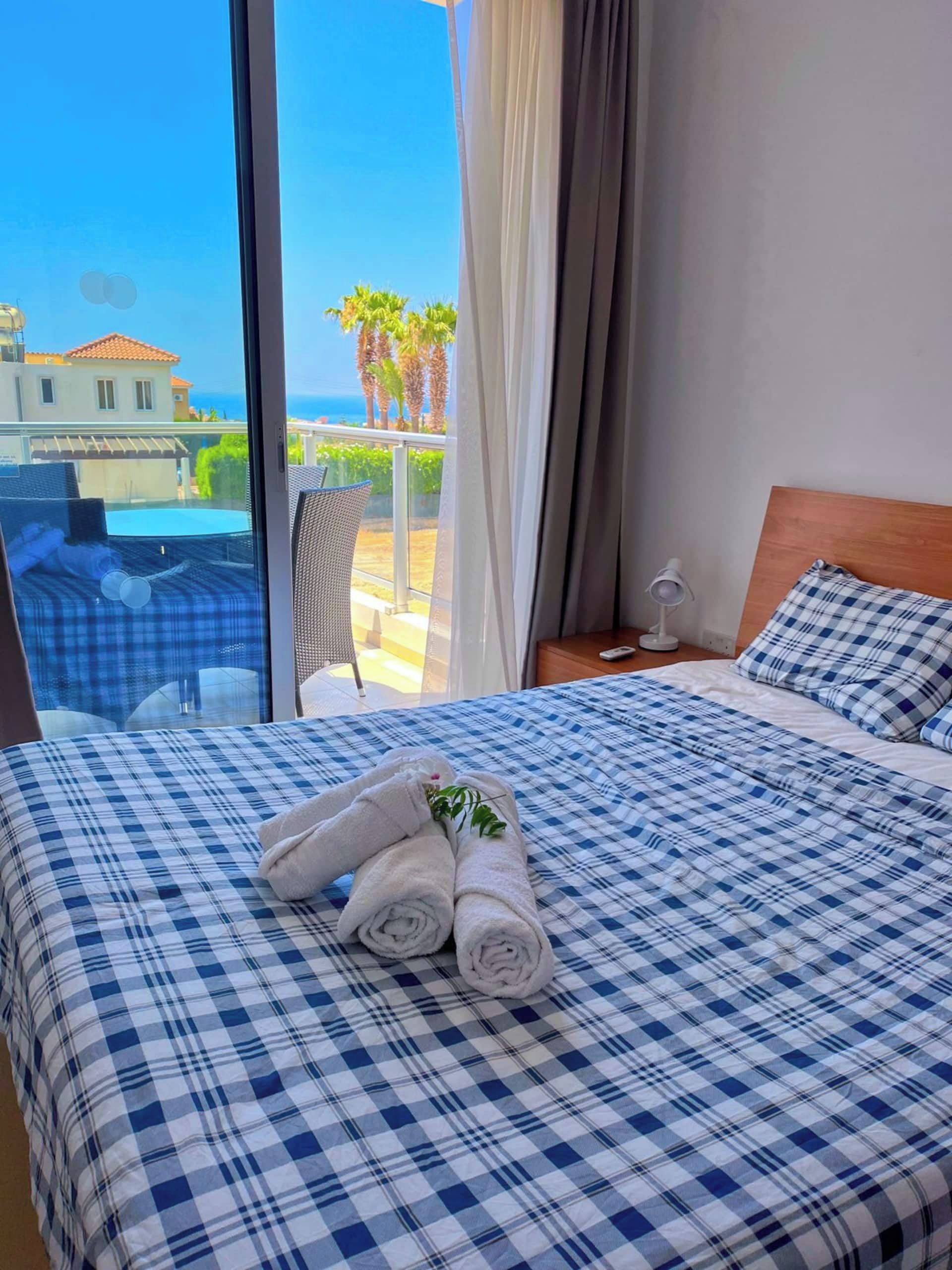 Une chambre d’hôtel avec une couvre-lit à carreaux, un lit bien fait avec des serviettes enroulées, une tête de lit en bois et une vue sur la mer et les palmiers, visible à travers de grandes fenêtres.