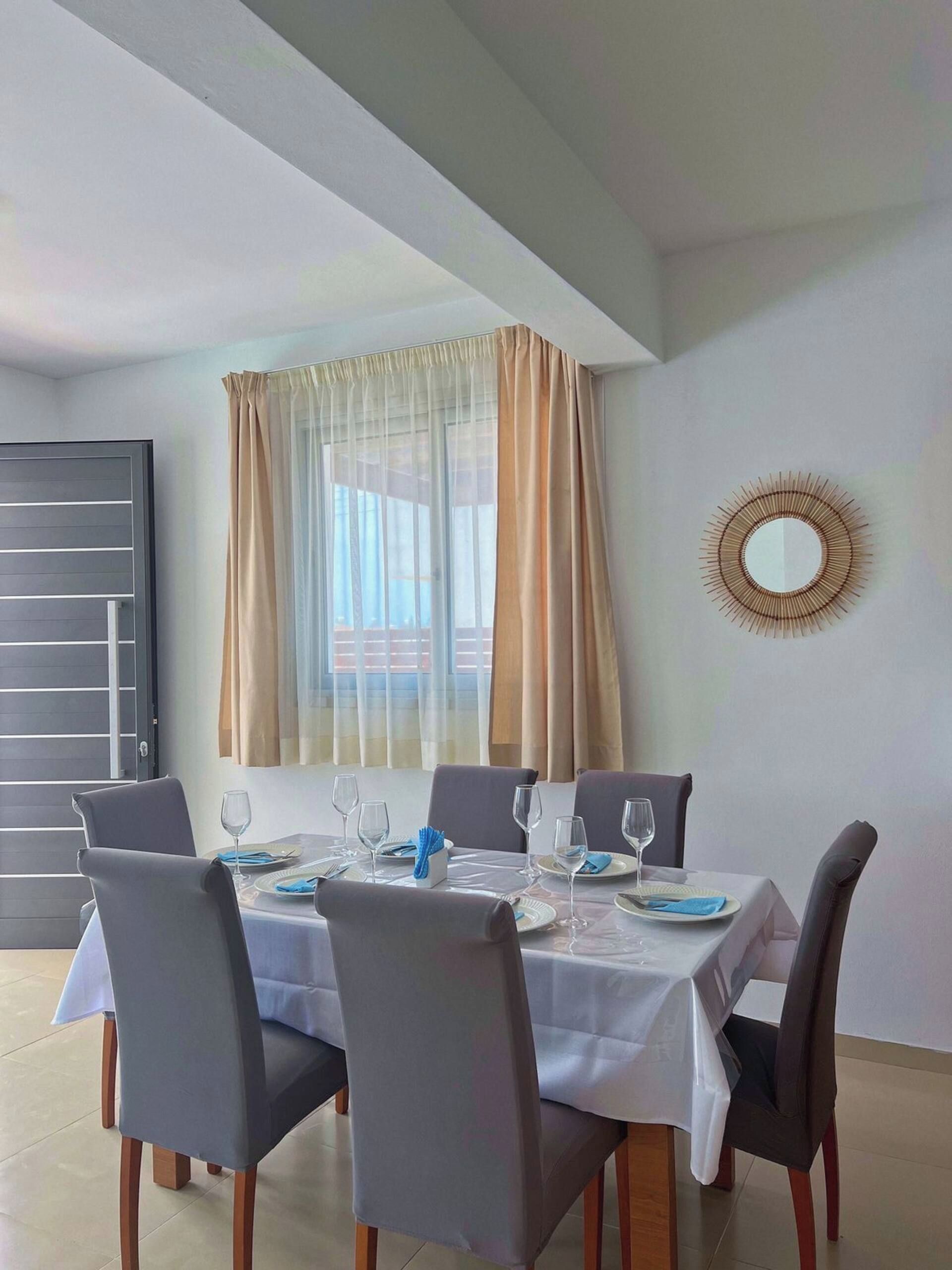 Un coin repas avec une table dressée pour six personnes, six chaises, une fenêtre avec des rideaux et un miroir fixé au mur.