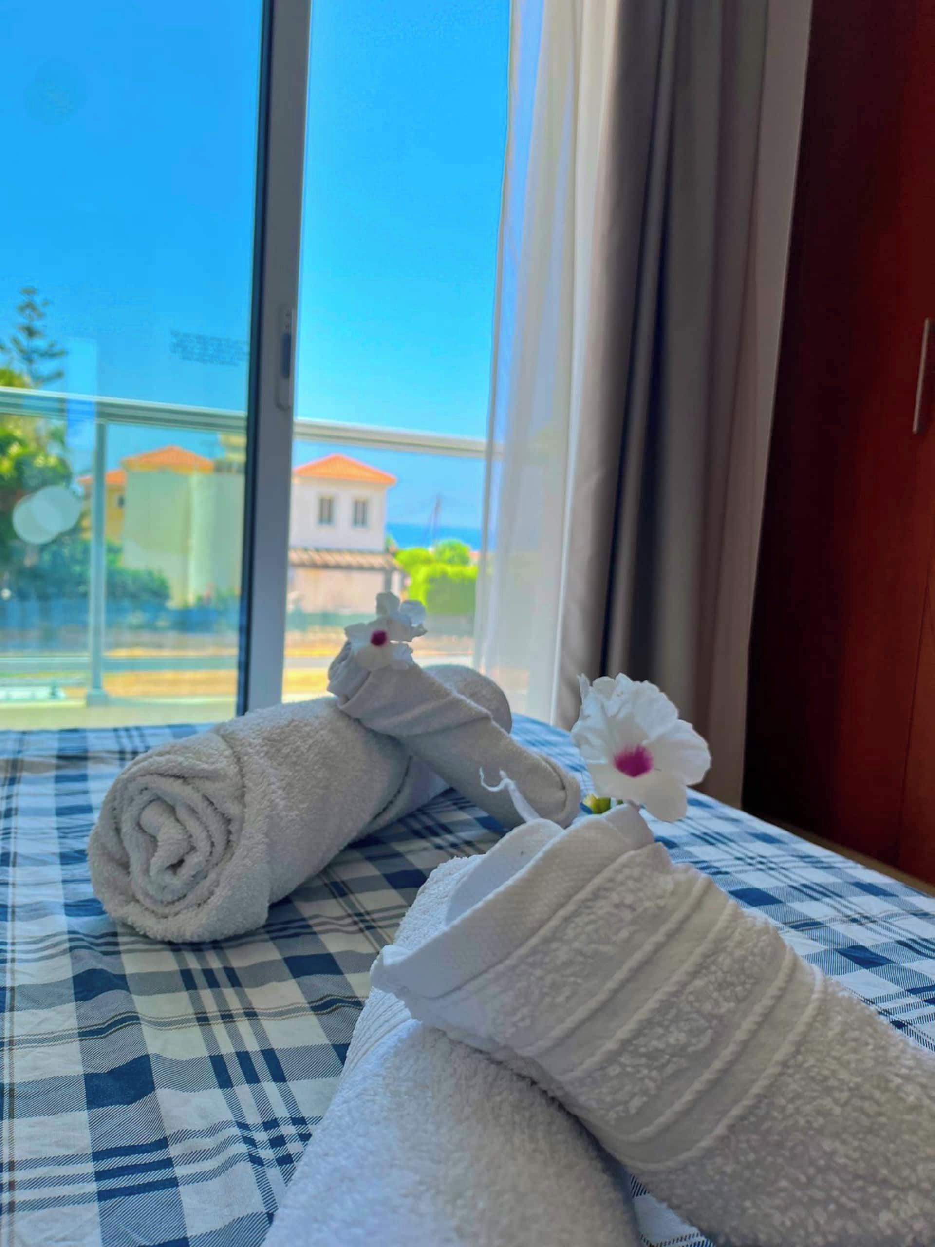 Ein Bett mit aufgerollten weißen Handtüchern und einer einzelnen weißen Blume auf einer karierten Bettdecke, mit Blick auf Gebäude und Grün durch ein Fenster.