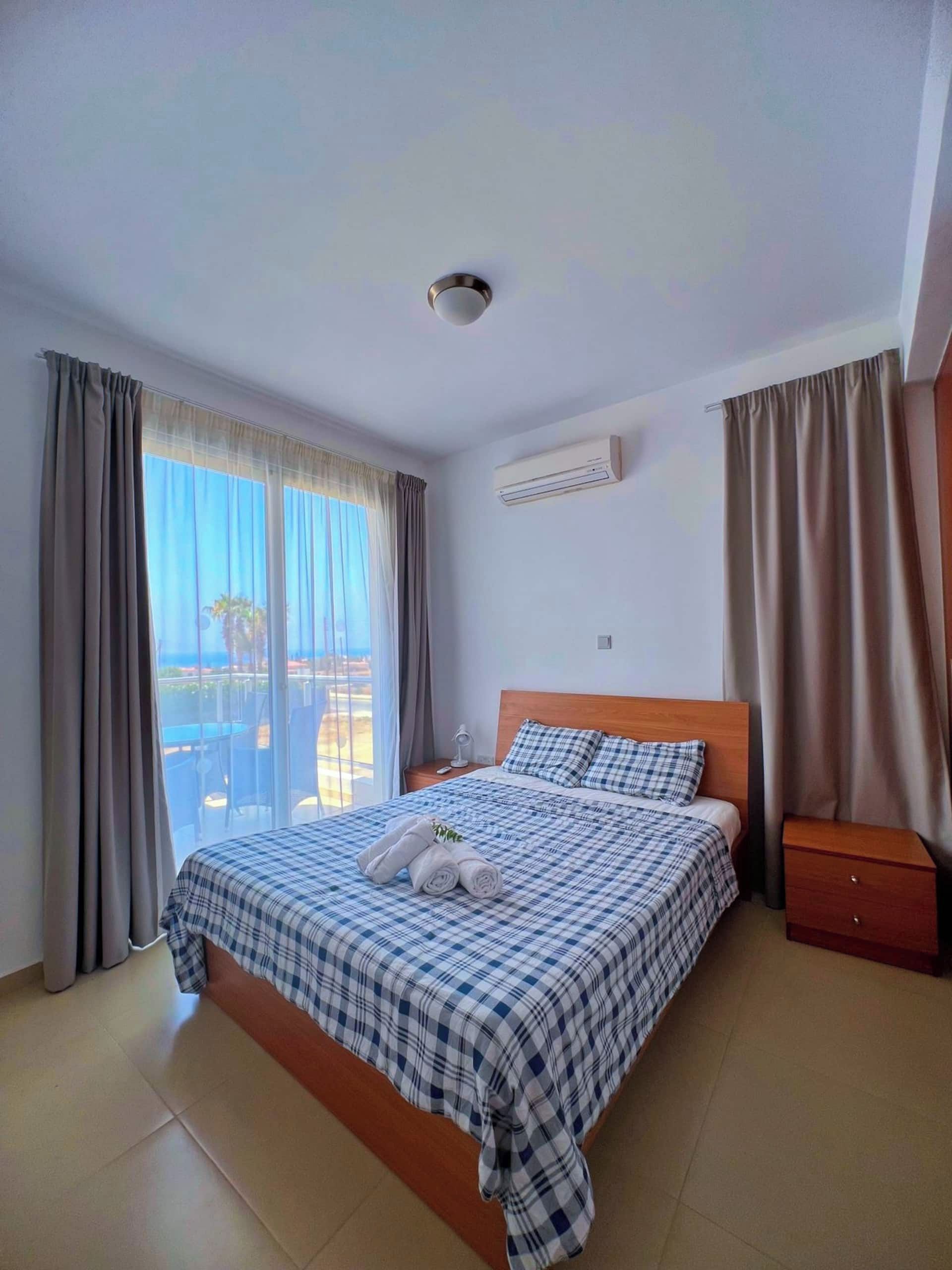 A hotel room with a bed, a nightstand, an air conditioning unit, and a view of the outdoors.