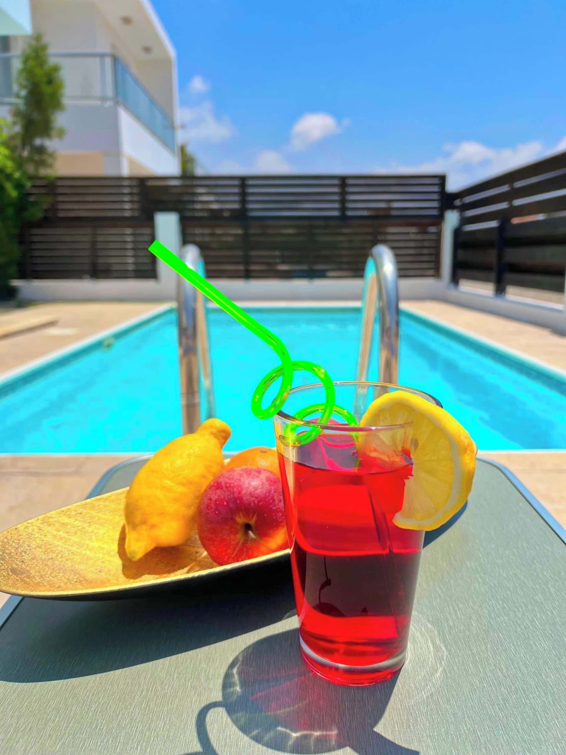 A poolside setting with a glass of red beverage, a plate with fruit, and a green straw.
