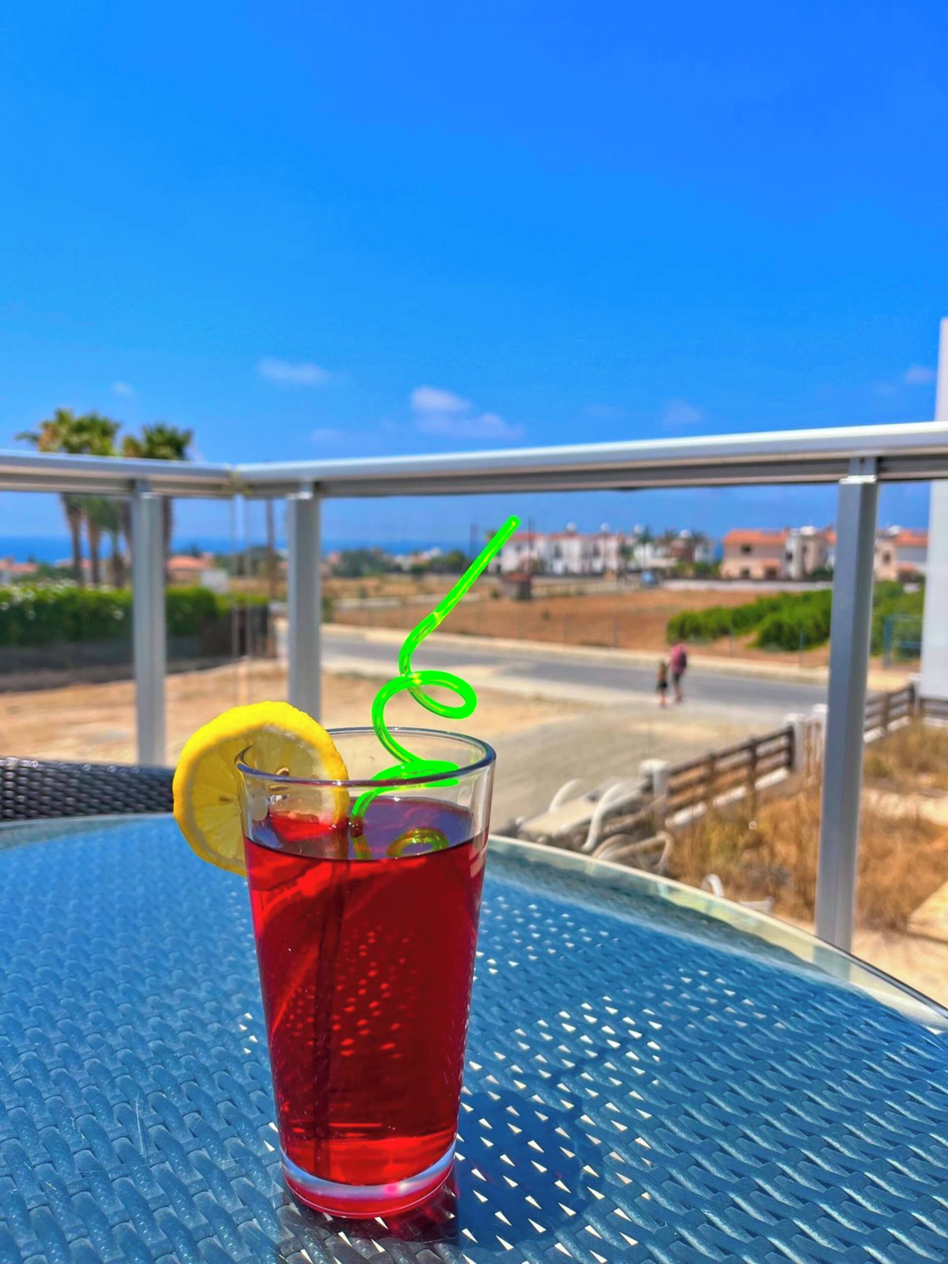 Un verre d’une boisson rouge, garni d’une tranche de citron et muni d’une paille verte, est posé sur une table en métal bleue.