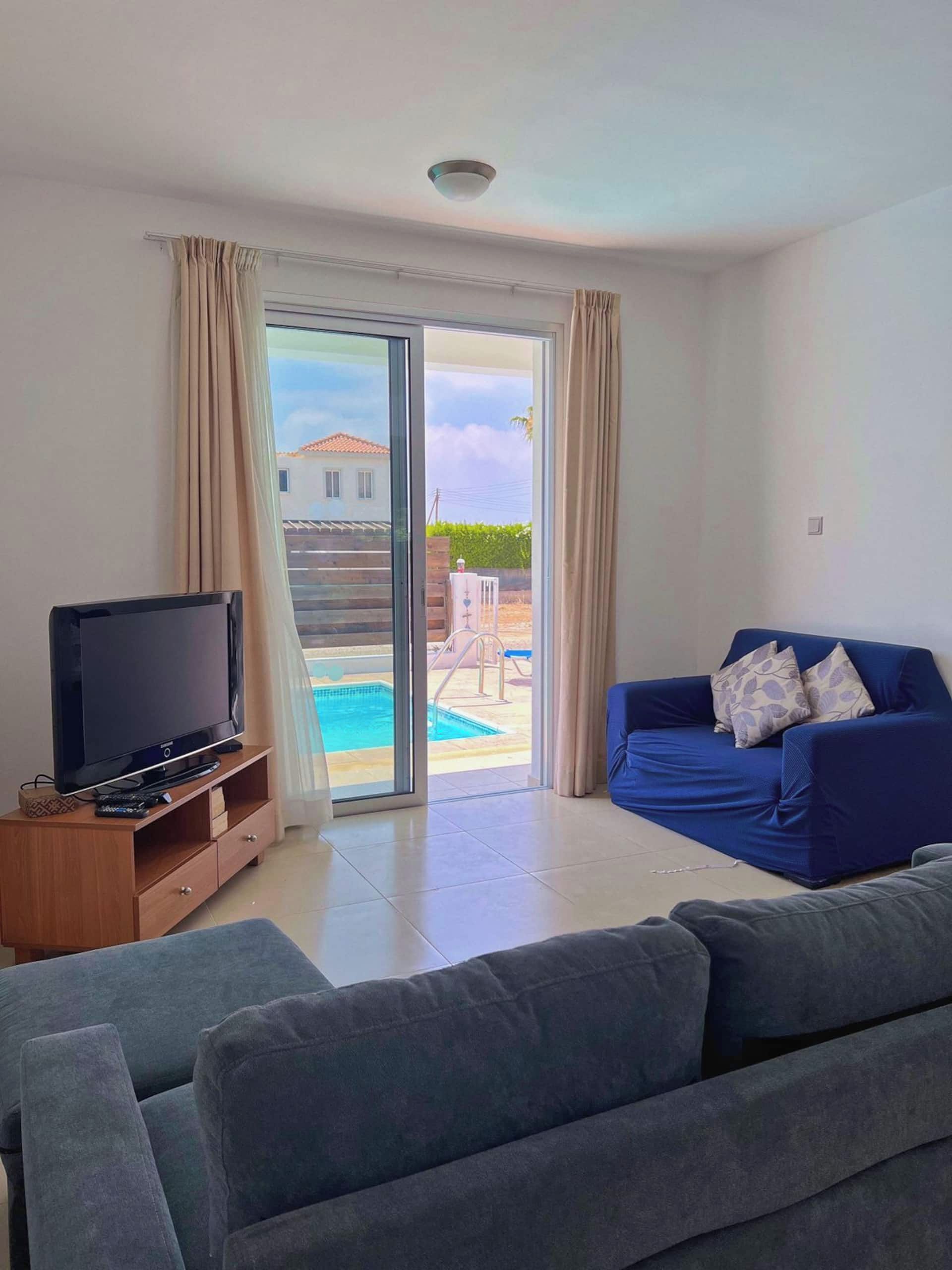 A living room with a TV, a sofa, and a view of a pool and house outside.