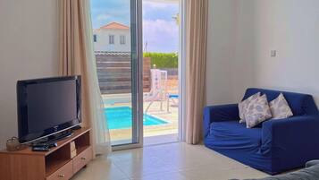 A living room with a TV, a sofa, and a view of a pool and house outside.