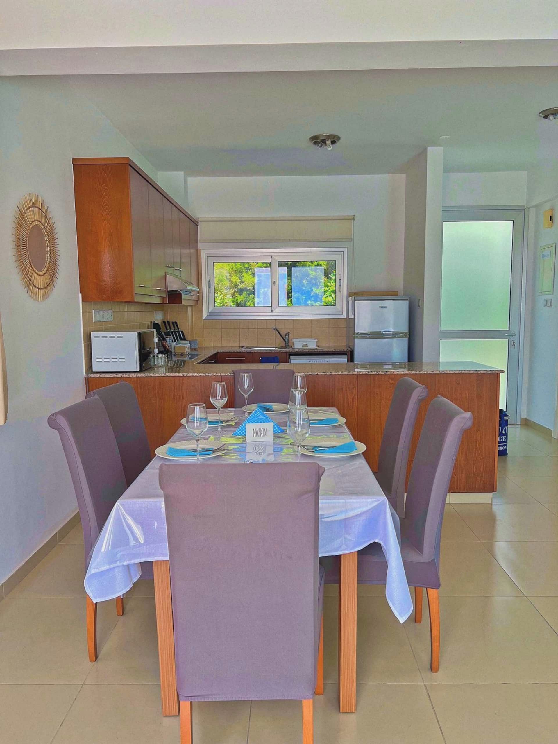 A dining area with a table set for four, a kitchen in the background, and a view of the outdoors through a window.