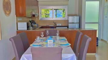 A dining area with a table set for four, a kitchen in the background, and a view of the outdoors through a window.